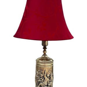 May include: A vintage table lamp with a red lampshade and a brass base. The base features a detailed relief of figures in a tavern scene. The lamp is a decorative lighting fixture.