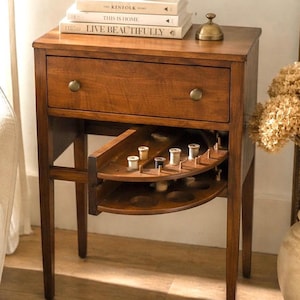 Puede incluir: Una mesa de costura de madera marrón cálido, con un cajón y una bandeja extraíble para guardar hilos. La mesa tiene pomos de latón y está coronada con libros y una taza de té. Ideal para coser y manualidades.