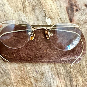 May include: Vintage gold-rimmed eyeglasses with clear lenses rest on a brown leather case. The glasses have a delicate, wire-frame design with a bridge and temples. The case is rectangular and shows signs of wear.