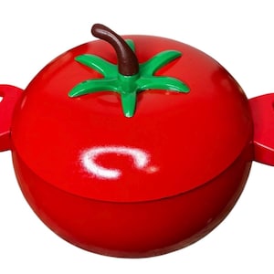 Puede incluir: Una olla redonda de color rojo brillante con dos asas laterales y una tapa a juego. La tapa presenta un tallo verde y una rama marrón curvada, diseñados para parecerse a un tomate. La superficie es lisa y brillante con un acabado reflectante.