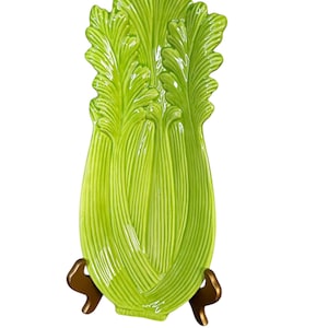 Puede incluir: Una fuente de cerámica con forma de tallo de apio, en un vibrante color verde. La fuente está sostenida por un soporte de metal dorado. El diseño presenta texturas detalladas de hojas y tallos, añadiendo un toque realista a la pieza decorativa.