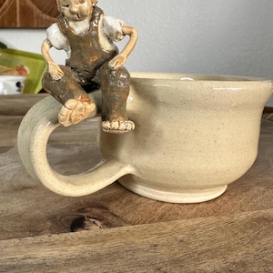 May include: A beige ceramic mug with a large handle, featuring a small figurine of a person sitting on the rim. The figurine is wearing brown overalls and a white shirt. The mug has a speckled texture.