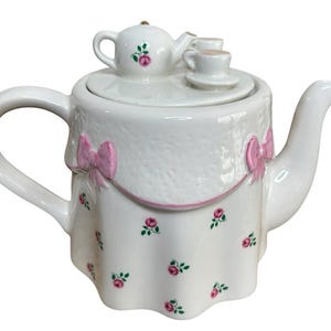 May include: A white ceramic teapot featuring a textured drape design with pink bows and a small floral rose pattern. The lid is topped with a miniature teapot and two tiny teacups on saucers, creating a decorative and layered appearance.
