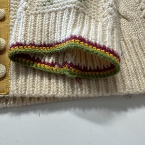 Op de afbeelding: Close-up van een cr&egrave;megekleurde gebreide mouw. De mouw heeft een geribbelde manchet met groene, bordeauxrode en gele randen. De trui heeft een ruitpatroon met blauwe, gele en rode accenten.