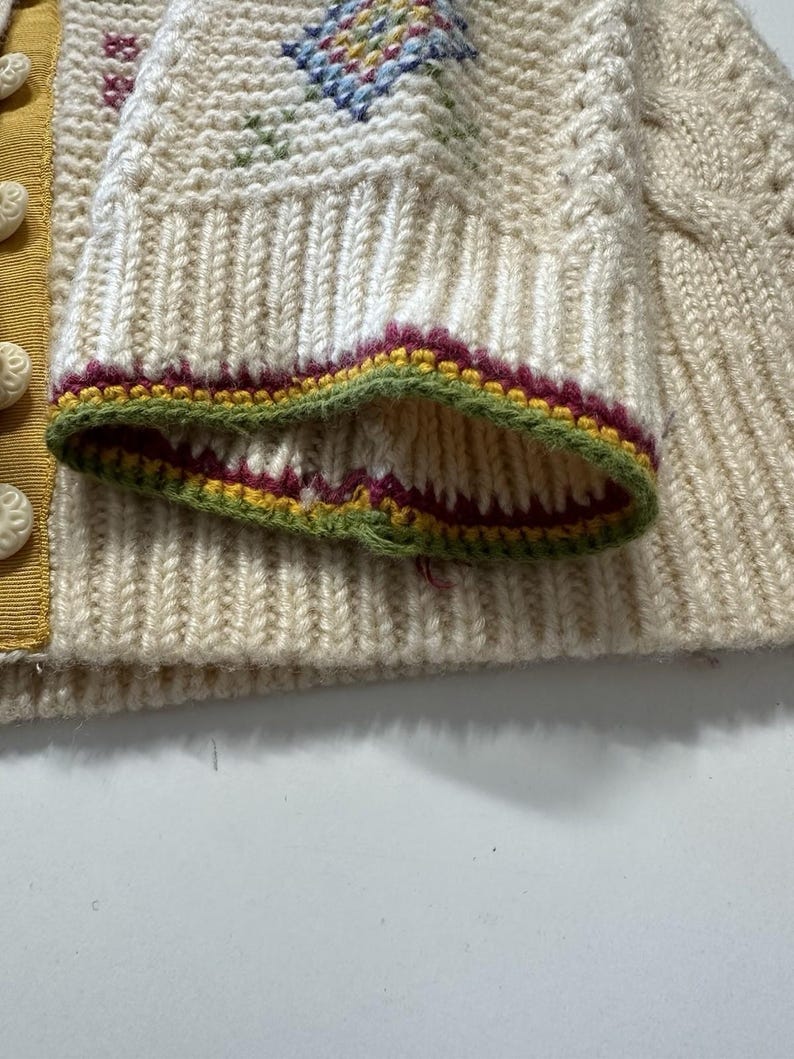 Op de afbeelding: Close-up van een cr&egrave;megekleurde gebreide mouw. De mouw heeft een geribbelde manchet met groene, bordeauxrode en gele randen. De trui heeft een ruitpatroon met blauwe, gele en rode accenten.