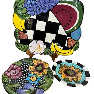 May include: A large rectangular ceramic platter with a black and white checkerboard center, decorated with painted grapes, watermelon, and bananas. Two smaller round ceramic plates feature colorful floral patterns with sunflowers and roses.