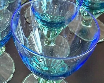 Könnte beinhalten: Ein Set aus durchscheinenden blauen Glasbechern mit einer breiten, abgerundeten Schale und einem dezenten Rippenmuster am Boden. Jeder Becher steht auf einem kurzen, dicken Stiel, der mit einem runden, klaren Glasfuß verbunden ist.