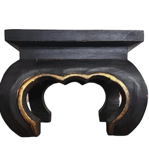 May include: A small, low-profile display stand featuring a flat, rectangular top surface supported by four curved legs. The stand is finished in a matte black color with gold-painted accents along the inner edges of the arched leg structure.