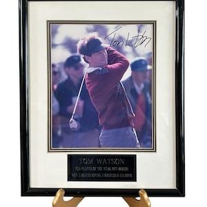 May include: Framed photograph of Tom Watson, a golfer, in action, with his signature. The frame is black with a white mat. A plaque below the photo reads "TOM WATSON" and lists his achievements. The frame is displayed on a gold-colored stand.