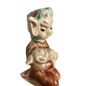 Op de afbeelding: Een keramieken beeldje van een knielende elfachtige figuur die een klein dier vasthoudt. Het beeldje heeft een bruin en beige kleurenschema met groene accenten op het hoofd en de nek. Het figuur heeft grote oren en een serene uitdrukking.