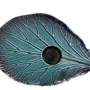 May include: A leaf-shaped ceramic dish in shades of teal and deep purple with a prominent vein pattern. The dish features a circular cutout in the center containing a black metal flower frog with numerous thin, upright pins for floral arrangement.
