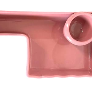May include: A pink ceramic serving dish with a unique shape. It features a rectangular tray with a small, round bowl attached. The dish is a solid, soft pink color and has a glossy finish.