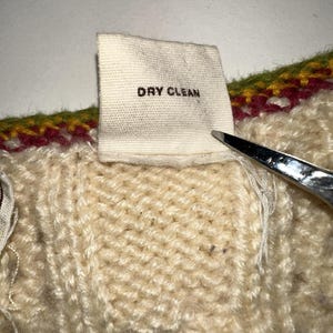 Op de afbeelding: Close-up van een cr&egrave;mekleurige gebreide stof met een label met de tekst "DRY CLEAN" in zwarte tekst. De stof heeft een getextureerd uiterlijk, met een kleurrijke rand van groen, rood en geel. Een schaar is gedeeltelijk zichtbaar.