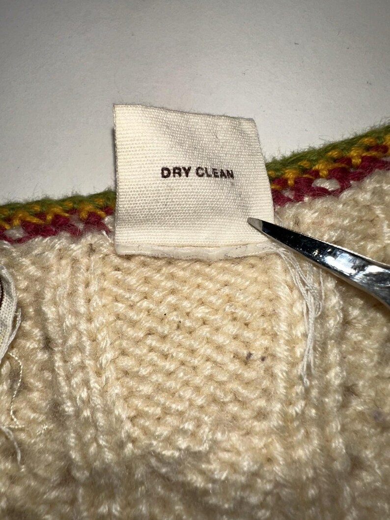 Op de afbeelding: Close-up van een cr&egrave;mekleurige gebreide stof met een label met de tekst "DRY CLEAN" in zwarte tekst. De stof heeft een getextureerd uiterlijk, met een kleurrijke rand van groen, rood en geel. Een schaar is gedeeltelijk zichtbaar.