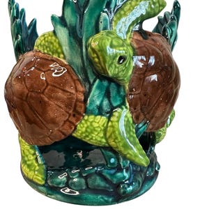 May include: A ceramic turtle figurine with a glossy finish. The figurine features two turtles with brown shells and green heads, surrounded by green and teal aquatic plants. The base is a dark teal colour.