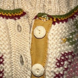 Op de afbeelding: Close-up van een cr&egrave;mekleurig gebreid vest met decoratieve bloemenborduurwerk. Het vest heeft een mosterdgele lintsluiting met twee witte knopen met bloemmotief. De halslijn is afgewerkt met groene, bordeauxrode en gele stiksels.