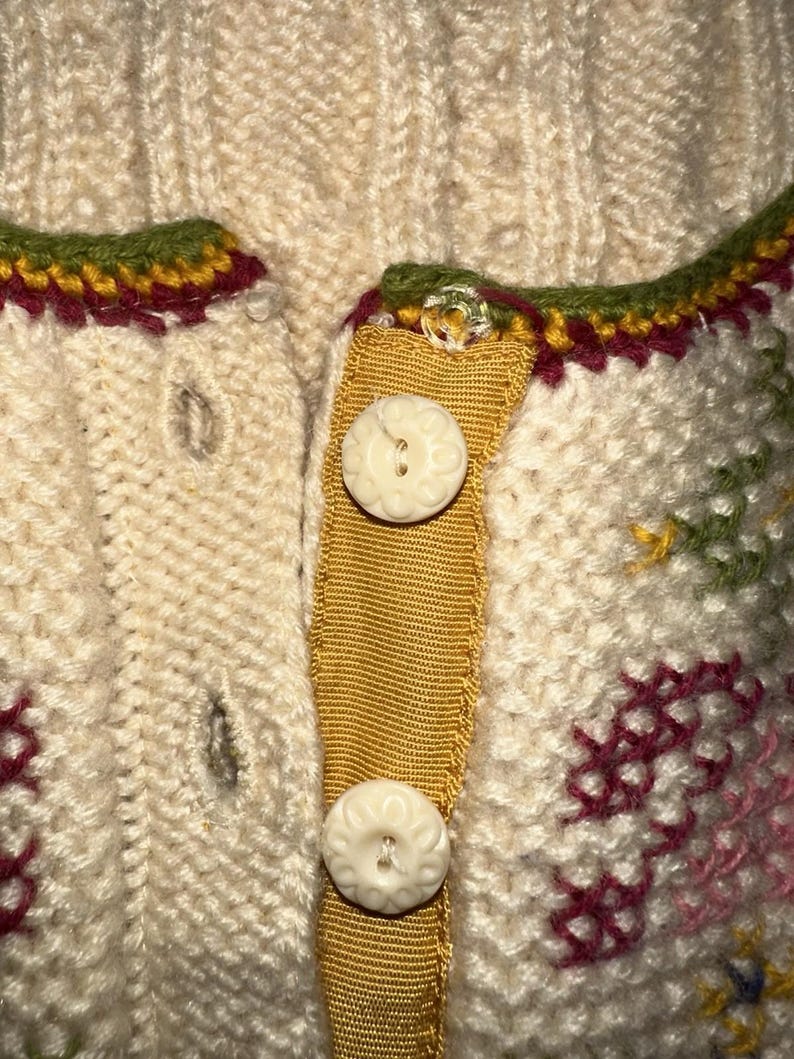 Op de afbeelding: Close-up van een cr&egrave;mekleurig gebreid vest met decoratieve bloemenborduurwerk. Het vest heeft een mosterdgele lintsluiting met twee witte knopen met bloemmotief. De halslijn is afgewerkt met groene, bordeauxrode en gele stiksels.