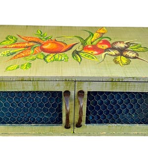 May include: A rectangular light green wooden cabinet with two doors featuring dark metal wire mesh panels and twisted metal handles. The top surface is decorated with a hand-painted illustration of orange carrots, red tomatoes, and dark root vegetables.