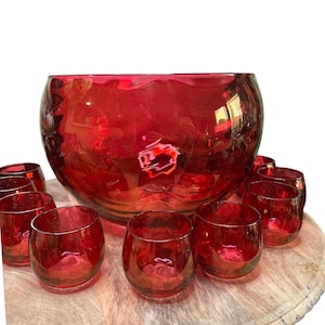May include: A red glass punch bowl set. The set includes a large, round punch bowl and several small, matching glasses. The glass has a glossy finish, and the set is displayed on a wooden serving tray. The color is a deep, rich red.