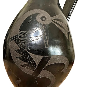 May include: A black ceramic pitcher with a handle and a wide base. The pitcher is decorated with an etched design of a bird-like figure with geometric shapes. The surface has a glossy finish.