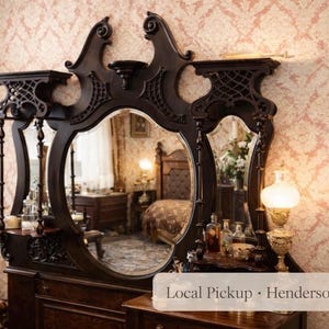 Op de afbeelding: Een donkerbruine antieke kaptafel met een grote ovale spiegel en twee kleinere spiegels. De kaptafel heeft sierlijke snijwerk en decoratieve planken. Een vintage lamp staat aan de zijkant, met behang met patronen op de achtergrond.