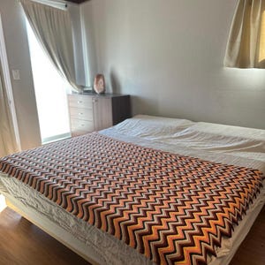 May include: A white bed with a brown, orange, and yellow chevron patterned crocheted throw blanket.