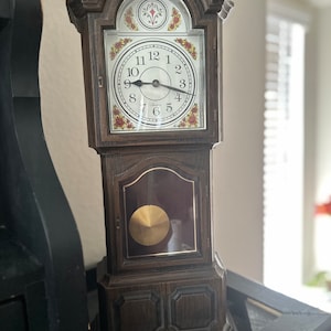 May include: A wooden grandfather clock with a white face and black hands. The clock has a gold pendulum and a decorative design on the front.