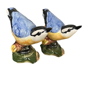 Puede incluir: Dos figuras de pájaros de cerámica. Cada pájaro tiene la espalda azul, la cabeza blanca y negra y el pecho amarillo. Están posados sobre una base marrón y verde. Las figuras son detalladas y parecen estar pintadas a mano.