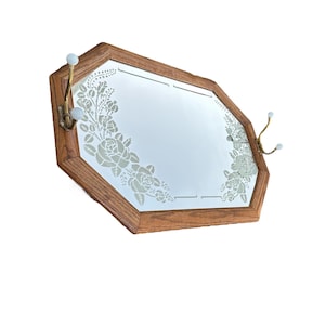 May include: Octagonal wooden framed mirror with etched floral design and two brass hooks with white ball ends. The mirror has a light brown wooden frame.