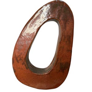 May include: A ceramic sculpture with a unique, open-center design. The artwork features a warm, reddish-brown color with darker, mottled accents. The glossy finish enhances the organic shape, creating a striking decorative piece.