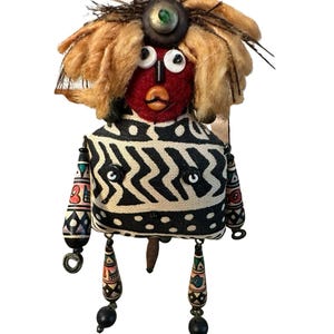 May include: A handcrafted doll with a red face, button eyes, and a button nose. The doll has a tan, fluffy hair-like design and a black and white patterned top. The doll's legs are adorned with colorful beads and dangle from the bottom.