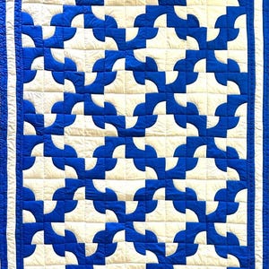 May include: A rectangular quilt featuring a repeating geometric pattern of interlocking blue and white curved shapes. The design is framed by a solid white border followed by a solid blue outer border, with visible grid-like stitching throughout.