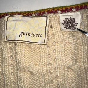 Op de afbeelding: Close-up van een cr&egrave;mekleurige gebreide trui met een textuurpatroon. De trui heeft een label met de tekst "guinevere" en een ander label met de samenstelling en wasvoorschriften. De bovenrand heeft een decoratieve rand met rode, gele en groene kleuren.