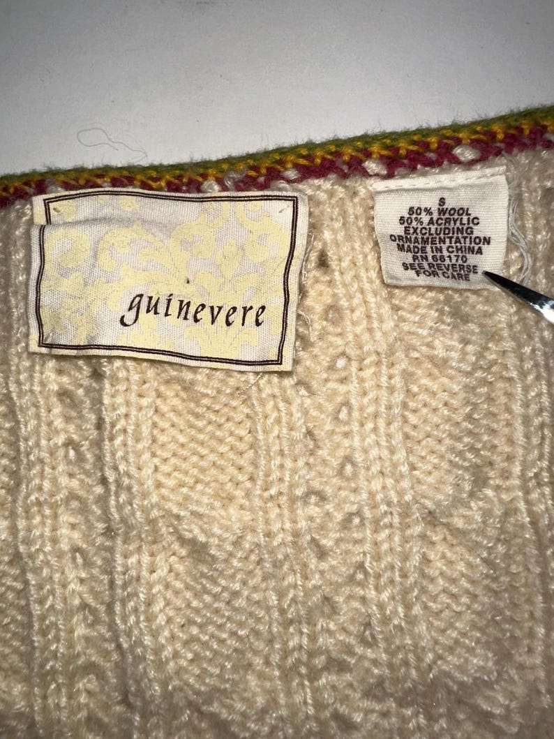 Op de afbeelding: Close-up van een cr&egrave;mekleurige gebreide trui met een textuurpatroon. De trui heeft een label met de tekst "guinevere" en een ander label met de samenstelling en wasvoorschriften. De bovenrand heeft een decoratieve rand met rode, gele en groene kleuren.