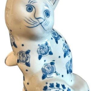 May include: A ceramic cat figurine in a seated pose, featuring a white base with blue floral and vine patterns. The cat's face has blue details, including eyes, whiskers, and ear accents. The figurine is a decorative item.