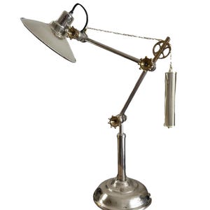 May include: A silver-toned desk lamp with a circular base and adjustable arm. The lamp features a round shade and decorative gears. A cylindrical weight hangs from a chain, adding to the industrial design.