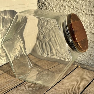 Pode incluir: Um frasco de armazenamento de vidro transparente com formato hexagonal e uma abertura inclinada. O frasco possui uma tampa circular de madeira castanho-escura que assenta num ângulo, proporcionando um fecho seguro para o recipiente.