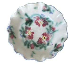 May include: A small, off-white ceramic dish with a ruffled edge and a hand-painted floral design. The flowers are primarily red with yellow centers, surrounded by green leaves and blue accents. The rim is outlined in blue.