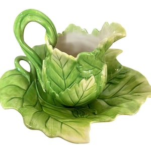 May include: A ceramic creamer and saucer set, designed to resemble a cabbage leaf. The creamer has a handle and is nestled on a matching leaf-shaped saucer. The set is painted in shades of green and white, creating a naturalistic appearance.
