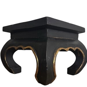 May include: A square black display stand featuring four curved legs with gold-colored accents along the inner edges. The stand has a flat top surface and a sturdy base, with a matte black finish covering the exterior surfaces of the structure.