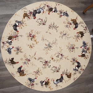 May include: A round, beige fabric tablecloth featuring a repeating pattern of roosters and hens interspersed with floral and wheat sprigs. The design is arranged in a circular motif across the surface, showcasing birds in black, brown, and red.