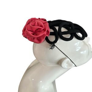 May include: A black headband with a series of connected loops and a large, red-pink fabric rose. The headband is displayed on a white mannequin head, showcasing the design and color contrast.