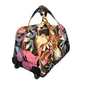 May include: A rolling travel bag with a floral pattern in pink, brown, white, and black. The bag has a pink zipper, black wheels, and a brown handle. The bag is designed for travel and has a rectangular shape.
