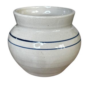 Könnte beinhalten: Ein weißer Keramiktopf mit rundem Körper und ausgestelltem Rand. Zwei horizontale blaue Streifen umkreisen den Topf. Der Topf hat ein rustikales, handgemachtes Aussehen, was darauf hindeutet, dass er zur Aufbewahrung oder Dekoration verwendet werden könnte.