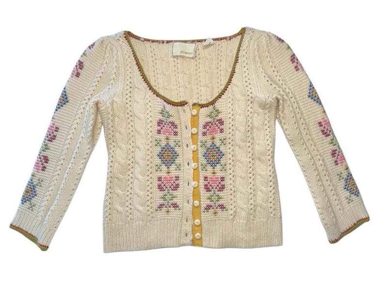 Op de afbeelding: Cr&egrave;mekleurig vest met kabelbreisel en bloemenkruissteekborduurwerk in roze, blauw en groen. De trui heeft een ronde hals, lange mouwen en een knoopsluiting met een gele bies. Afmetingen in centimeters.