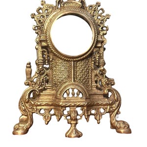 May include: A gold-toned metal decorative stand with an ornate design. The stand has a round top with a raised edge and three legs. The stand is intricately detailed with scrollwork and floral motifs.