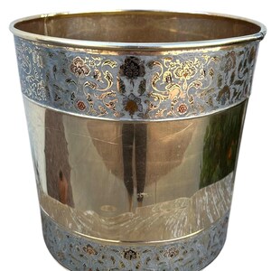 Peut inclure: Une corbeille à papier cylindrique en métal avec une bande centrale dorée polie et des motifs floraux gravés complexes dans des tons argentés et dorés sur les bords supérieur et inférieur, offrant un aspect réfléchissant et décoratif.