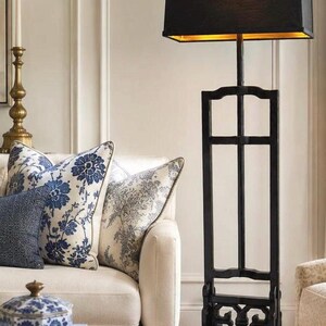 May include: A tall floor lamp with a black rectangular frame and a matching black rectangular lampshade. The interior of the lampshade is gold. The base of the lamp features a decorative openwork design with a central cross-shaped support structure.