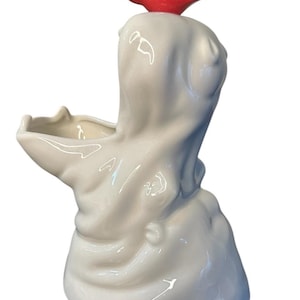 Puede incluir: Una maceta de cerámica blanca con forma de pájaro, con una pequeña abertura para plantas. Una figura de pájaro rojo brillante se encuentra en la parte superior. La maceta tiene un acabado brillante y está diseñada para contener plantas o flores pequeñas.