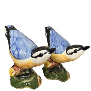 Könnte beinhalten: Zwei Keramikvogel-Figuren. Jeder Vogel hat einen blauen Rücken, einen weißen und schwarzen Kopf und eine gelbe Brust. Sie sitzen auf einem braunen und grünen Sockel. Die Vögel blicken in die gleiche Richtung.